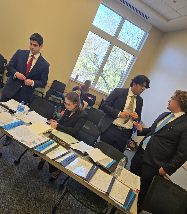 Group of students dressed in suits and business attire standing around a table with papers on it preparing for trial.